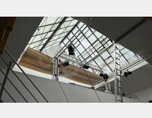Innenansicht eines modernen Raumes mit einem geneigten Glasdach, das Tageslicht hereinlässt. Im Vordergrund befindet sich eine Metallkonstruktion mit Scheinwerfern und Lautsprechern, die offenbar für Beleuchtungs- und Tonzwecke verwendet wird.