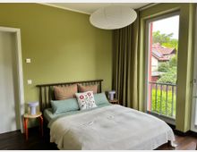 Schlafzimmer mit olivgrünen Wänden, Doppelbett mit dunkel gerahmtem Kopfteil und zwei kleinen Nachttischen. Rechts führt eine große Glastür mit Vorhang auf einen Balkon und bietet Blick auf Bäume und benachbarte Häuserdächer.