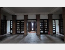 Raum mit bodentiefen Glastüren und mehreren Säulen aus braunem Stein, die den Eingang zu einer Tür mit horizontalen Glaselementen in der Mitte betonen. Der Fußboden besteht aus Steinplatten, und an der rechten Seite steht ein Feuerlöscher.