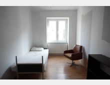 Schmales, schlichtes Schlafzimmer mit weißen Wänden und hellem Holzfußboden, einem Einzelbett an der linken Wand und einem braunen Sessel. An der Stirnwand befindet sich ein großes Fenster mit Heizkörper darunter; rechts steht ein dunkles Regal/Schrankmöbel.