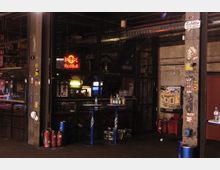 Industrieähnlicher Innenraum einer Bar oder eines Clubs mit dunklem, robustem Design, Gitterelementen und metallischen Oberflächen. Im Vordergrund stehen blaue Stehtische, umgeben von Getränkeflaschen, mit einem Neon-Red-Bull-Logo und verschiedenen Wandgraffitis sowie Aufklebern im Hintergrund.