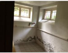 Ein kleiner Kellerraum mit zwei vergitterten Fenstern, abblätterndem Putz an den Wänden und sichtbaren Wasserrohren. Der Boden ist mit hellen Fliesen ausgelegt.