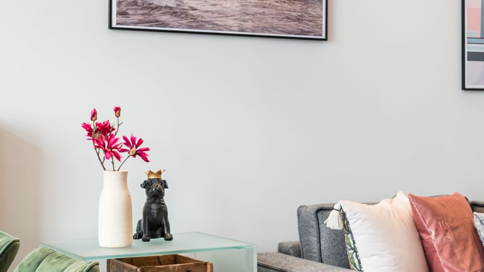 Heller Wohn- und Essbereich mit grauem Sofa und farbigen Kissen, daneben ein kleiner Beistelltisch mit Vase und Dekoobjekt. Im Vordergrund stehen grüne Polsterstühle am Holztisch; an der weißen Wand hängen gerahmte Fotografien.