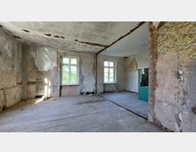 Ein leerer, verfallener Raum mit abblätterndem Putz an den Wänden, freiliegendem Mauerwerk und sichtbaren Schäden an Decke und Boden. Zwei große Fenster lassen Tageslicht herein, und die Wandtapete ist an einigen Stellen abgerissen.