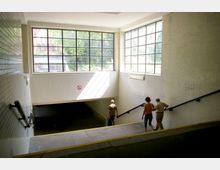 Ein unterirdischer Treppenabgang mit weißen, gefliesten Wänden und großen Fenstern, durch die Tageslicht einfällt. Drei Personen steigen die Treppe hinab, während ein Schild mit Rauchverbot an der Wand sichtbar ist.