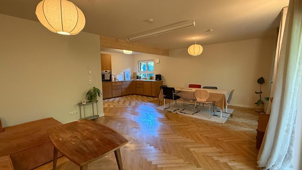 Ein offener Wohn- und Essbereich mit Holzfußboden in Fischgrätmuster, einer zentralen Esstischgruppe mit verschiedenen Stühlen und einer modernen Küche im Hintergrund. Zwei runde, hängende Lampen und bodenlange Vorhänge an den Fenstern verleihen dem Raum eine helle Atmosphäre.