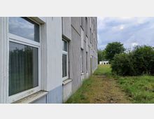 Außenansicht eines grauen, mehrstöckigen Gebäudes mit rechteckigen Fenstern, flankiert von einer Wiese und Büschen. Im Hintergrund sind ein kleineres weißes Gebäude und Bäume sichtbar, unter einem bewölkten Himmel.