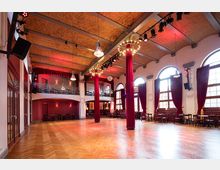 Großer Veranstaltungssaal mit hölzernem Fischgrätparkettboden, hohen Decken und roten Samtvorhängen an den großen, bogenförmigen Fenstern. Markante Säulen mit roten Stoffverkleidungen und Kronleuchtern sowie eine erhöhte Empore im Hintergrund.