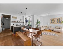 Offener Wohnraum mit moderner Küche, Essbereich und Wohnzimmer. Der Raum verfügt über Holzböden, eine schwarze Kücheninsel mit Barhockern, einen Holztisch mit Stühlen, eine beige Eckcouch und Pflanzen, mit viel natürlichem Licht durch große Fenster.