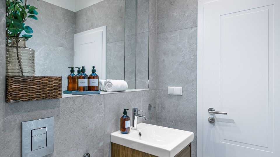 Modernes, kleines Badezimmer mit grauen Wand- und Bodenfliesen, wandhängendem Waschbeckenunterschrank aus Holz und großem Spiegel mit Schienenbeleuchtung. Links sind ein Unterputz-Spülkasten, Toilettenpapierhalter und Ablage mit Pflanze und Flaschen zu sehen, rechts eine weiße Tür.