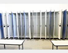 Reihen von halb geöffneten Umkleideschränken aus Metall in einem hellen Umkleideraum mit gefliesten Böden; im Vordergrund steht eine schlichte Sitzbank.