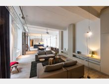 Großzügiger Raum in modernem Loft-Stil mit offener Gestaltung, hellen Wänden und großen Fenstern. Im Vordergrund befindet sich eine Sitzecke mit grauen Sofas, im Hintergrund ein langer Esstisch und eine minimalistische Küche mit Hängelampen.