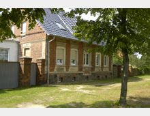 Backsteinhaus mit Satteldach und weißen Fensterrahmen, teilweise restauriert, umgeben von einer Wiese und Bäumen. Links ist ein Metallzaun mit Backsteinpfeilern zu sehen.