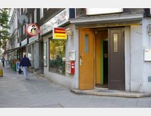 Straßenansicht mit einer kleinen Sportsbar und Geschäften an einem Gehweg in einer Stadthäuserzeile. Sichtbar sind geöffnete Eingangstüren, Werbeschilder, ein Schaufenster mit Pflanzen und ein vorbeilaufender Passant.