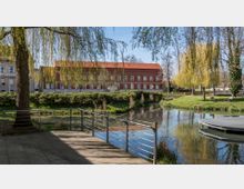 Ein Park mit einem kleinen Gewässer, im Vordergrund ein gepflasterter Bereich mit Metallgeländer und einer Baumkrone, im Hintergrund ein rotes, zweistöckiges Gebäude mit vielen Fenstern und rotem Ziegeldach, umgeben von Bäumen und gepflegten Grünflächen.