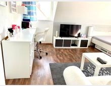 Helles, modernes Schlafzimmer unter einer Schräge mit Holzfußboden. Ein weißer Schreibtisch mit einem Drehstuhl und Dekorationsgegenständen steht links, während sich ein TV auf einem weißen Lowboard rechts befindet; ein Bett und ein Couchtisch ergänzen den Raum.