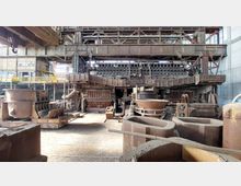 Industrielle Produktionshalle mit einer großen, rostigen Stahlkonstruktion, mehreren Gussgefäßen und Maschinen. Die Umgebung zeigt metallene Strukturen, Rohre und Geländer sowie hohe Fenster, die Tageslicht in den Raum lassen.