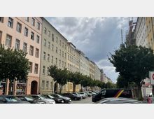 Straßenszene mit einer Reihe von mehrstöckigen Wohngebäuden in pastellfarbenem Putz, flankiert von Bäumen entlang der Straße. Im Vordergrund sind geparkte Autos und ein dunkler Lieferwagen zu sehen, während am rechten Bildrand ein Gebäude mit Gerüsten steht.