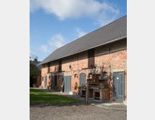 Backstein-Gebäude im ländlichen Stil mit grauen Holztüren und kleinen Fenstern; davor stehen rustikale Holzmöbel und Pflanzen. Der Hof ist teils mit Kies und Rasen bedeckt, mit blauem Himmel im Hintergrund.