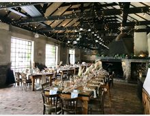 Ein rustikaler Veranstaltungsraum mit Holzbalkendecke, hängender Lichterkettendekoration und großen, fensterartigen Glasflächen. Im Raum stehen gedeckte Holztische mit Stühlen, Dekorationen aus Blumen und Pampasgras, und im Hintergrund ist ein großer Steinkamin sichtbar.