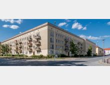 Wohnblock im Stil der Nachkriegsarchitektur mit hellgelber Fassade, mehreren Stockwerken und Balkonen. Das Gebäude steht an einer gepflasterten Straßenecke, umgeben von Bäumen, vereinzeltem Grün und Straßenlampen unter einem blauen Himmel mit wenigen Wolken.