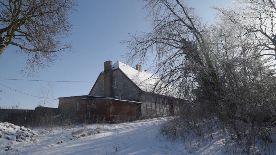 Winterliche ländliche Hofstelle mit einem großen, älteren Wohnhaus mit schneebedecktem Satteldach und einem niedrigen Backsteinanbau. Ein verschneiter Weg führt am Gebäude vorbei, umgeben von kahlen Bäumen und Sträuchern unter klarem Himmel.