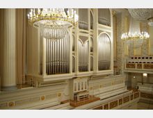 Innenraum eines Konzertsaals mit großer klassischer Orgel, umrahmt von weißen und goldenen Verzierungen. Im Vordergrund sind ein Podium mit einer Tastatur sowie glänzende Kristalllüster und gepolsterte Sitzreihen sichtbar.