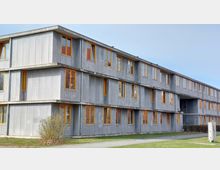 Ein modernes, rechteckiges Gebäude mit grauer Fassade und zahlreichen Fenstern mit orangen Rahmen. Es hat drei Stockwerke, einen Gehweg davor und ist von einer Rasenfläche mit Sträuchern umgeben.