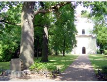 Eine kleine Kirche mit weißer Fassade steht in einem von Bäumen umgebenen Park. Ein gepflasterter Weg führt durch den gepflegten Rasen direkt zum Eingang der Kirche, während im Vordergrund ein Gedenkstein unter einem großen Baum sichtbar ist.