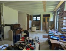 Renovierungsraum mit teilweise sichtbarer Backsteinwand, Holzbalkendecke und großen Fenstern auf der rechten Seite. Im Raum befinden sich Werkzeuge, ein Klavier, ein Kaminrohr, ein Tisch mit Schüsseln und Baumaterialien, sowie eine Sitzecke mit einem Sessel im Hintergrund.