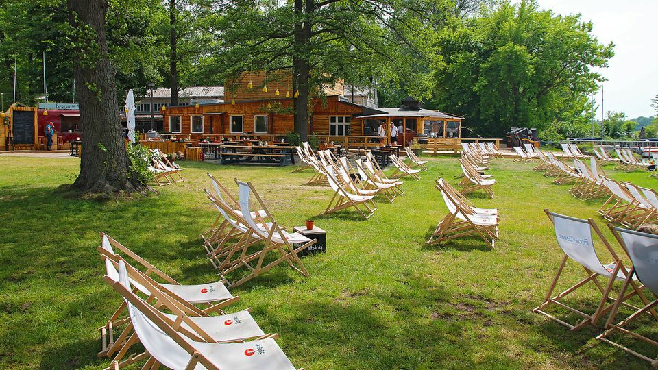 Grüne Außenanlage mit Holzgebäuden im Hintergrund, umgeben von Bäumen. Auf der Rasenfläche stehen zahlreiche Liegestühle und Holztische, die auf einen gastronomischen Bereich hinweisen.