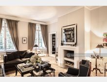 Helles Wohnzimmer mit Parkettboden, großen Fenstern mit beigen Vorhängen und einem weißen Kamin. Im Raum stehen schwarze Polstermöbel, ein Couchtisch mit Büchern und Blumen sowie ein Beistelltisch mit Lampen und Dekorationen.