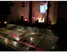 Ein Veranstaltungssaal mit einer großen Audiomischkonsole im Vordergrund. Im Hintergrund ist eine Bühne mit einer projizierten Aufnahme eines Fotos und mehreren sitzenden Zuschauern im gedimmten Licht zu sehen.