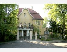 Gelbes zweistöckiges Wohnhaus im Stil des Historismus mit Ziegeldach, geschwungener Giebelfassade und mehreren verzierten Fenstern; ein schmiedeeisernes Tor führt zu einem Vorgarten. Umgeben von Bäumen, befindet sich das Gebäude in einer ruhigen, grünen Umgebung.