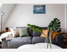 Wohnzimmer mit grauem Sofa, dekoriert mit bunten Kissen und einer gelben Decke, im Hintergrund ein abstraktes Wandbild und helle Wände. Im Vordergrund steht ein runder Couchtisch mit einer Vase, die große grüne Blätter enthält.