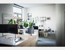 Helles, modernes Büro mit mehreren weißen Schreibtischen und schwarzen Bürostühlen. Auf den Tischen stehen Computerbildschirme, Pflanzen und verschiedene Arbeitsmaterialien; im Hintergrund sind eine große Fensterfront und an der Wand Bilder und Dekorationen zu sehen.