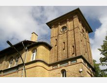 Backsteingebäude mit einem hohen Turm und dekorativen Zierelementen aus farbigen Kacheln, großen Rundbogenfenstern, und einem flachen Dach; im Vordergrund eine Straßenlaterne, umgeben von teilweise bewölktem Himmel.