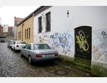 Gepflasterte Seitenstraße mit geparkten Autos neben Gebäuden mit verputzten und gelben Backsteinfassaden, die Wände sind mit Graffiti bedeckt. Im Vordergrund ist eine dunkelbraune Tür in einer weißen Hauswand zu sehen.