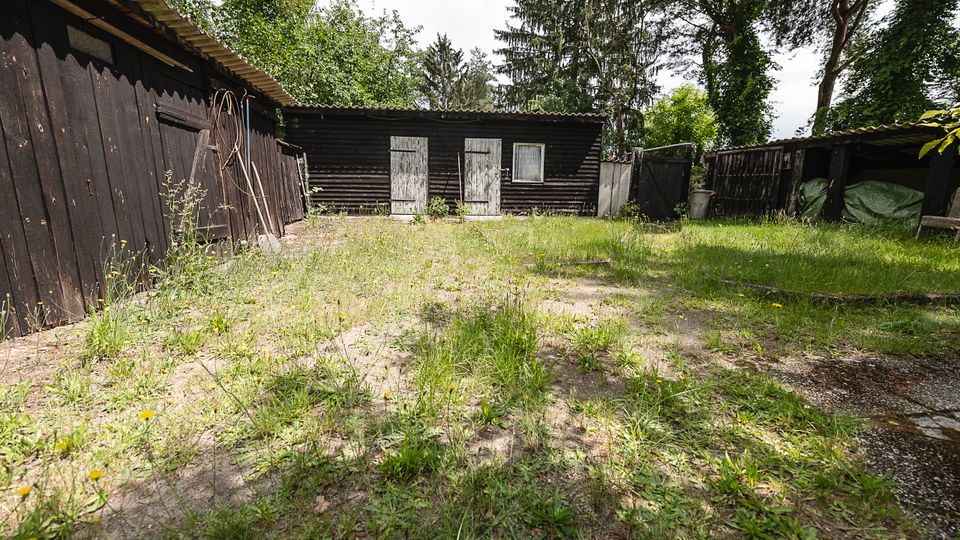 Innenhof mit unebenem, teils sandigem Boden und spärlichem Grasbewuchs, umgeben von dunklen Holzschuppen mit Wellblechdächern. Im Hintergrund stehen zwei helle Holztüren und ein kleines Fenster, daneben Zäune und hohe Bäume.