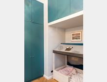 Blaugrüne Einbauschränke in einem Kinderzimmer, kombiniert mit einem weißen, hölzernen Hochbett, das mit einem kleinen Stoffdach und bunter Bettwäsche ausgestattet ist; an der weißen Wand hängt ein gerahmtes Bild.