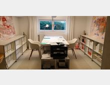 Ein helles Büro mit zwei weißen Sesseln an einem Tisch, der mit Unterlagen bedeckt ist. Im Hintergrund befindet sich ein Fenster mit Vorhängen, davor ein Heizkörper, und an den Wänden stehen Regale mit Aktenordnern und Dokumenten; ein Drucker steht auf einem kleinen Regal im Vordergrund.