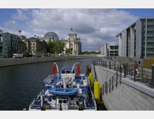 Ein Flussufer in einer Stadt mit einem modernen Regierungsviertel im Hintergrund, darunter der Reichstag mit seiner gläsernen Kuppel. Im Vordergrund liegt ein Boot mit blauer Lackierung und roten Sonnenschirmen an einer Anlegestelle.