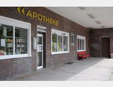 Außenansicht einer Apotheke mit einer Fassade aus dunklem Klinkerstein, großen Schaufenstern und einem Glaseingang. Über dem Eingang befindet sich ein gelbes Schild mit der Aufschrift „APOTHEKE“, daneben steht eine rote Bank unter einem überdachten Gehweg.