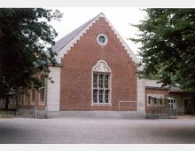 Backsteinbau mit Satteldach und einem großen, verzierten Fenster in der Mitte der Fassade. Das Gebäude hat helle Steinverzierungen an den Ecken, ein kleines Rundfenster im Giebel und steht auf einem Kiesplatz, umgeben von Laubbäumen.