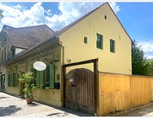 Gelbes Gebäude mit Satteldach, grünen Fensterläden und einem dekorativen Schild mit ovaler Beschriftung an der Fassade. Rechts schließt ein großer Holztor-Zaun den Hof ab; im Vordergrund Gehweg mit Pflastersteinen, sonnig beleuchtet.