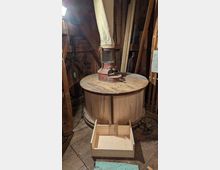 Innenraum einer historischen Mühle mit einem großen runden Holzmahlwerk und einem zentralen Maschinenaufsatz mit Stoffschlauch. Rundherum stehen Holzbalken, Werkzeugteile und Bretter, auf dem Holzboden liegt vorne eine helle Holzkiste.
