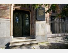 Eingangsbereich einer Synagoge mit verzierten Metalltüren, umgeben von einer Fassade aus Backstein und Stein. Rechts befindet sich eine Gedenktafel mit hebräischem Stern sowie ein schmiedeeisernes Geländer, das den Bereich abgrenzt.