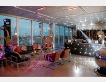 Modern eingerichteter Veranstaltungsraum mit großen Panoramafenstern, die eine städtische Skyline bei Sonnenuntergang zeigen. Der Raum ist mit Lounge-Möbeln, einer Bar, dekorativen Pflanzenelementen und einer Discokugel beleuchtet, die Lichteffekte im Raum erzeugt.