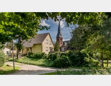 Ein gelbes Wohnhaus mit Satteldach steht in einer grünen, von Bäumen und Büschen umgebenen Umgebung. Im Hintergrund ist eine rote Backsteinkirche mit hohem Kirchturm und spitzem Dach zu sehen, während ein Gehweg und ein Wegweiser im Vordergrund sichtbar sind.