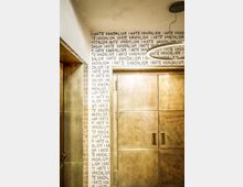 Innenansicht eines Flurs oder Eingangsbereichs mit großen, metallischen Doppeltüren und einer runden Deckenleuchte. Die Wand ist über mehrere Reihen mit dem wiederholten Schriftzug „I HATE VANDALISM“ in schwarzer Handschrift bedeckt.